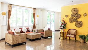 Un vestíbulo amplio con área de descanso que incluye sofás de mimbre y una silla de madera, ventanas grandes con cortinas y una pared de acento amarilla con detalles decorativos.