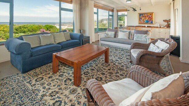 A spacious living room with a large window offering a view of the sea, a blue sofa, a wooden coffee table, and wicker furniture.