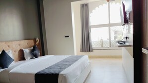 Habitación de hotel con cama, ventana con cortinas, televisor montado en la pared y una mesita con toma de corriente.