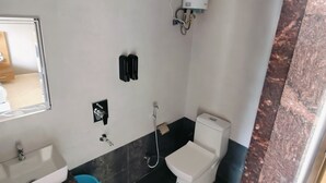 Un baño con lavamanos, inodoro y ducha blancos. Se observa un balde azul y un calentador de agua blanco montado en la pared.
