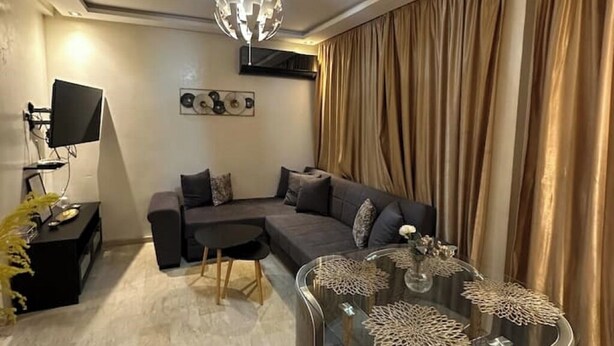 Uma sala de estar moderna com um sofá seccional, uma mesa de centro de vidro com objetos decorativos, uma televisão de tela plana e um candelabro.