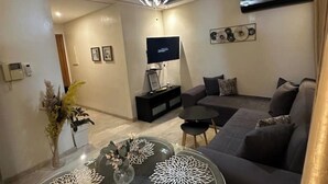 Uma sala de estar moderna com um sofá cinzento, uma mesa de centro com objetos decorativos, uma TV em um suporte preto e um luminário decorativo.