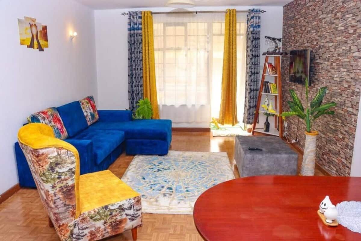Een woonkamer met een blauwe bank, een gestreept fauteuil, een ronde houten tafel en een boekenkast.