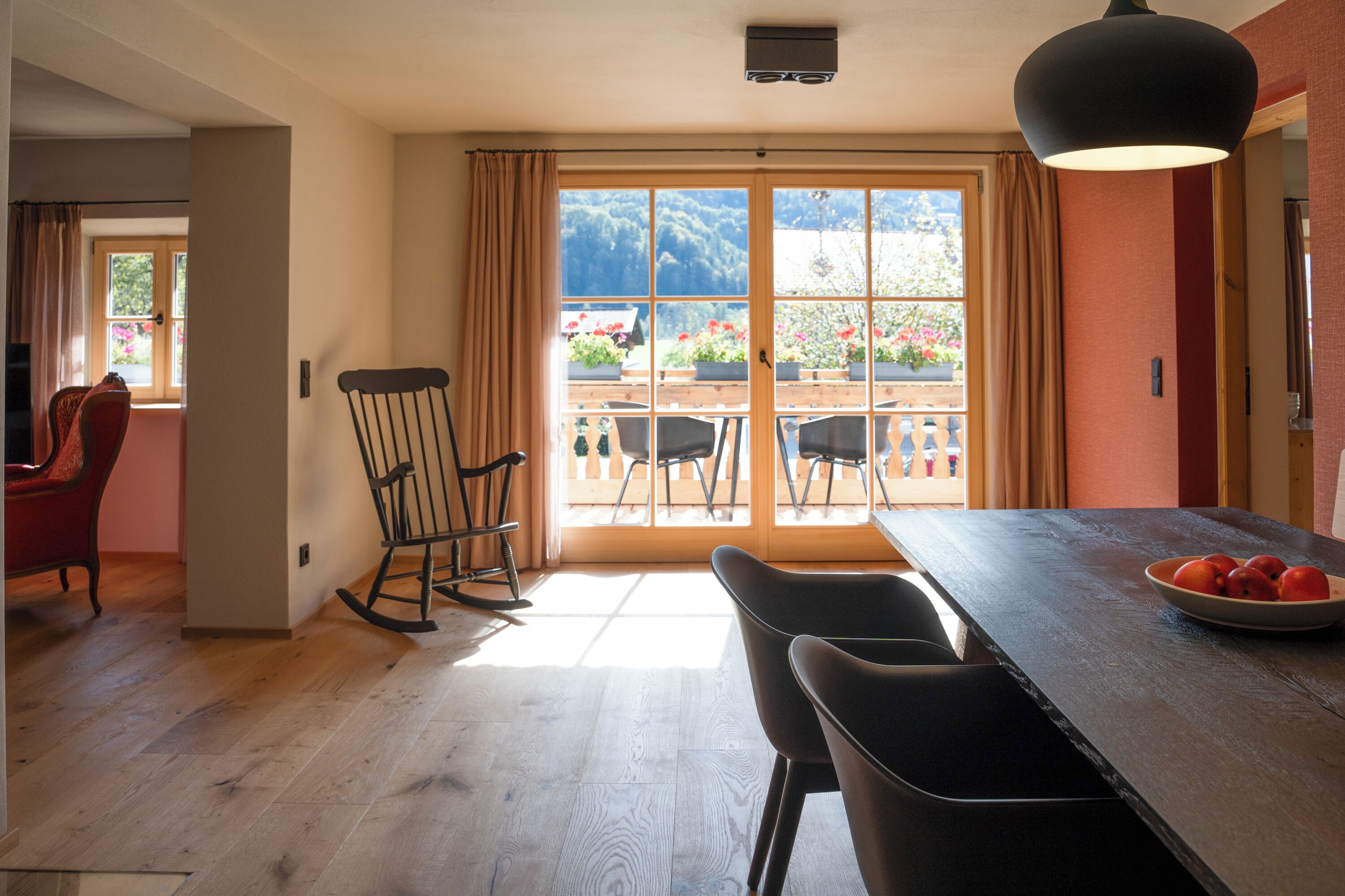 A dining area with a wooden table, black chairs, a rocking chair, and a view of a garden through a glass door.