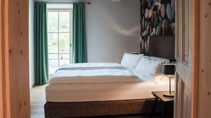 A bedroom with a large bed, a bedside table with a lamp, and a view of the outdoors through a window with green curtains.