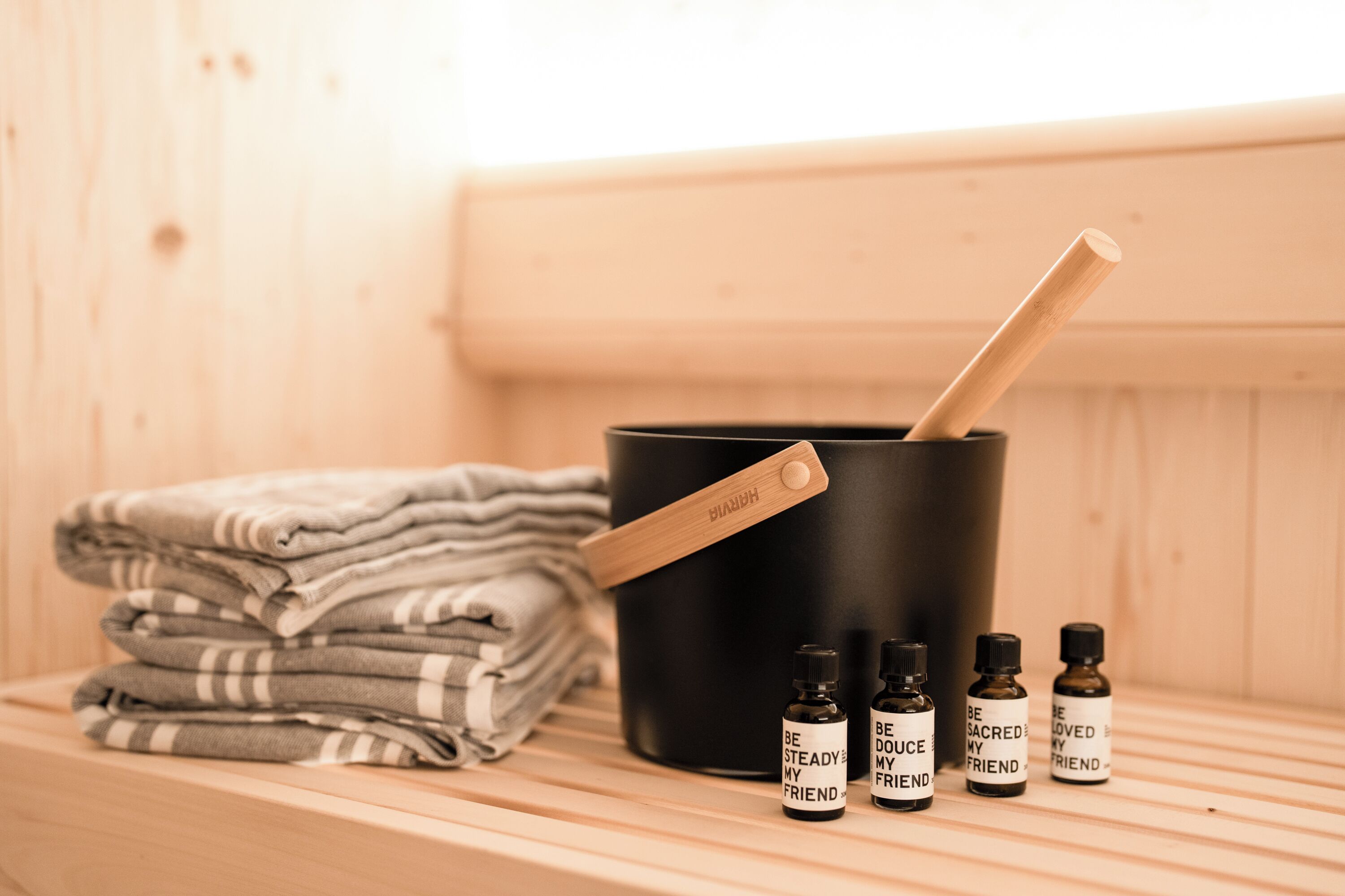 Una sauna con un banco de madera, un recipiente negro con una cuchara de madera y tres botellas pequeñas con etiquetas.