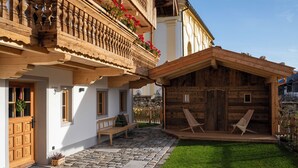 A traditional wooden chalet with a balcony adorned with red flowers, a stone-paved courtyard, and a grassy lawn.