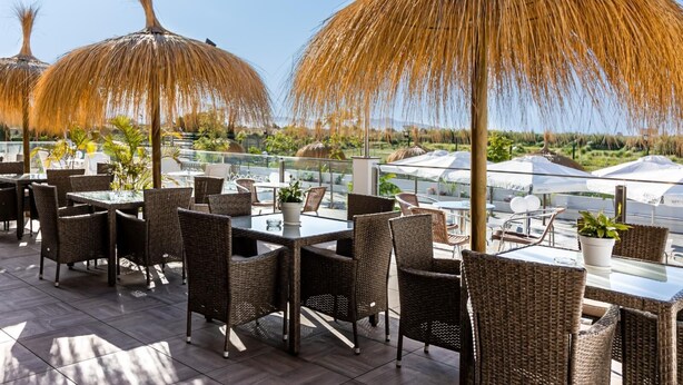 An outdoor dining area with wicker furniture and thatched umbrellas, overlooking a scenic landscape.