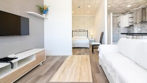 A modern living room with a flat-screen TV, a white sofa, a wooden coffee table, and a bed in the background.