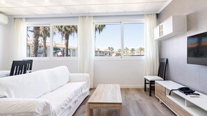 A modern, well-lit room with a white sofa, a wooden coffee table, a TV mounted on the wall, and large windows offering a view of palm trees and buildings.