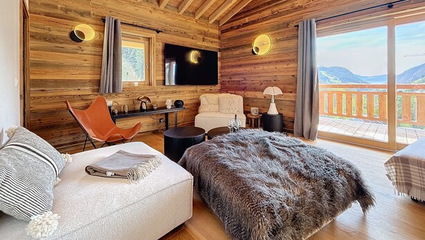A cozy cabin interior with a wooden ceiling, a large TV, a sofa, a chair, a small table, and a view of the outdoors. - Clos d'arsene - Les Gets - Proche centre (Les Gets)