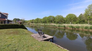 A calm river with a dock and a chair, surrounded by greenery and a building. - Bungalow in Zuytland Buiten by the Water (Simonshaven)
