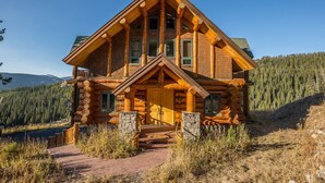 A log cabin with a prominent front porch, stone pillars, and a view of a forested mountain range.