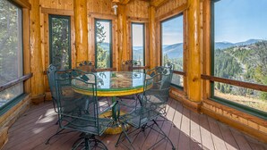 A wooden cabin with a glass-top dining table surrounded by metal chairs, large windows offering mountain views, and a wooden deck.