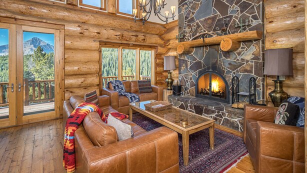 A log cabin interior with a stone fireplace, leather sofas, and a large window overlooking a forested mountain.