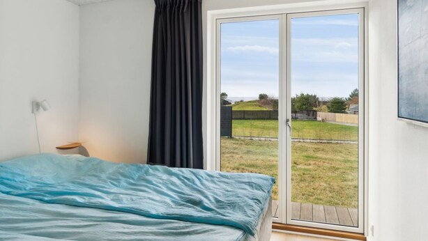 Ein Schlafzimmer mit einem Bett, einem Nachttisch, einem Fenster mit Vorhängen und Blick auf eine grüne Fläche.