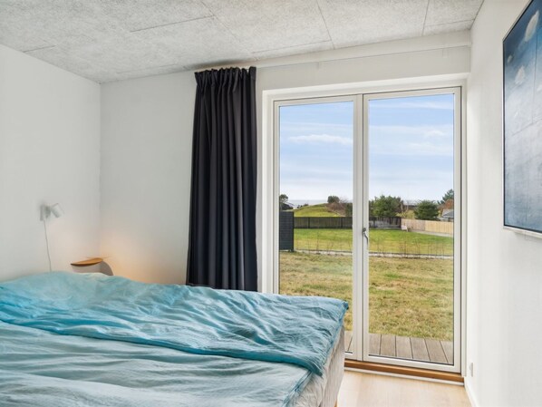 A bedroom with a bed, a nightstand, a window with curtains, and a view of a grassy area outside.