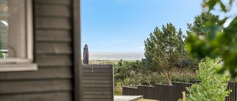 Une vue depuis une fenêtre donnant sur une cour verdoyante, avec une terrasse en bois, un arbre et une étendue d’eau au loin.