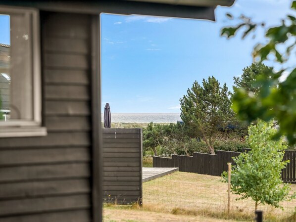 A view from a window overlooking a grassy yard with a wooden deck, a tree, and a distant body of water.