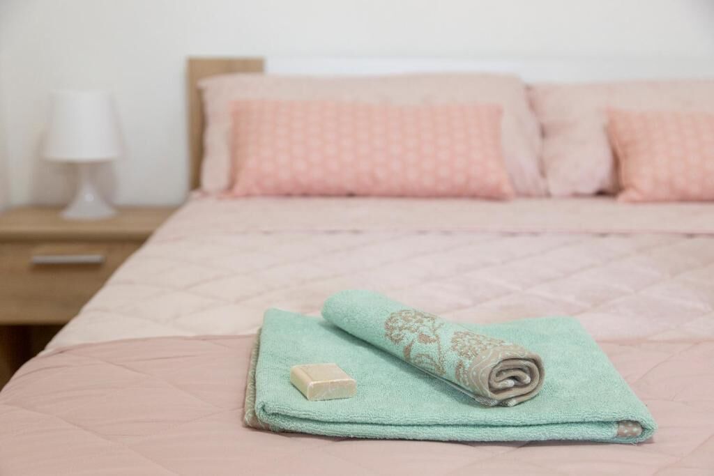 Cama bem arrumada com colcha e travesseiros cor-de-rosa, toalha enrolada e uma barra de sabão sobre a cama.