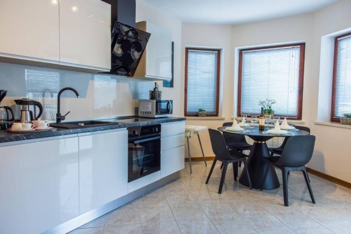 A modern kitchen with white cabinets, a built-in oven, a microwave, and a coffee maker. There is a dining table with chairs and a window with blinds.