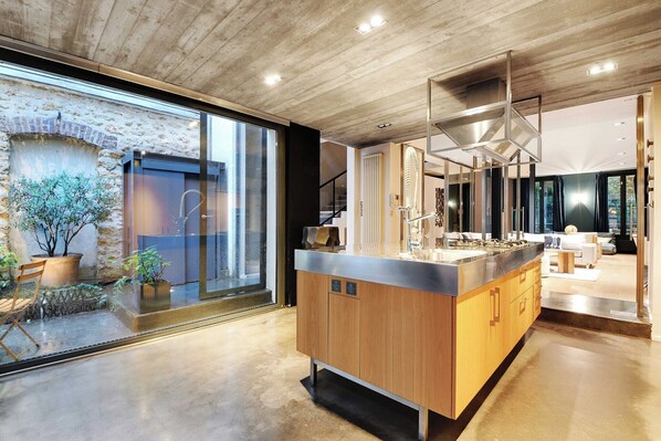 A modern kitchen with a large island, wooden cabinets, and a view of a garden through large windows.