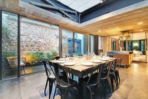 A modern dining area with a large table set for six, surrounded by black chairs, with a stone wall and greenery visible through large windows.