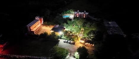An aerial view of a well-lit area with a large building, a swimming pool, and a parking lot.