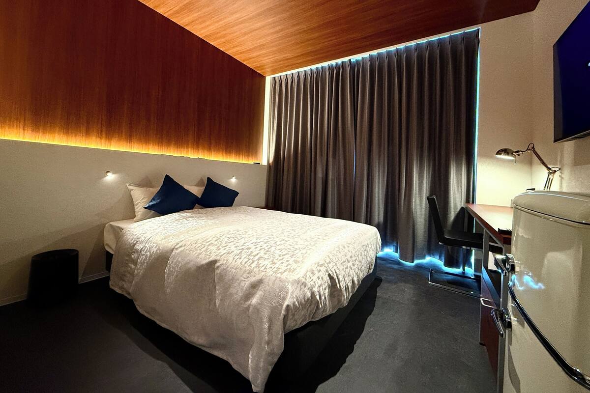 Ein modernes Hotelzimmer mit einem großen Bett, dunklen Vorhängen, einem Schreibtisch mit Lampe und einem an der Wand befestigten Fernseher.