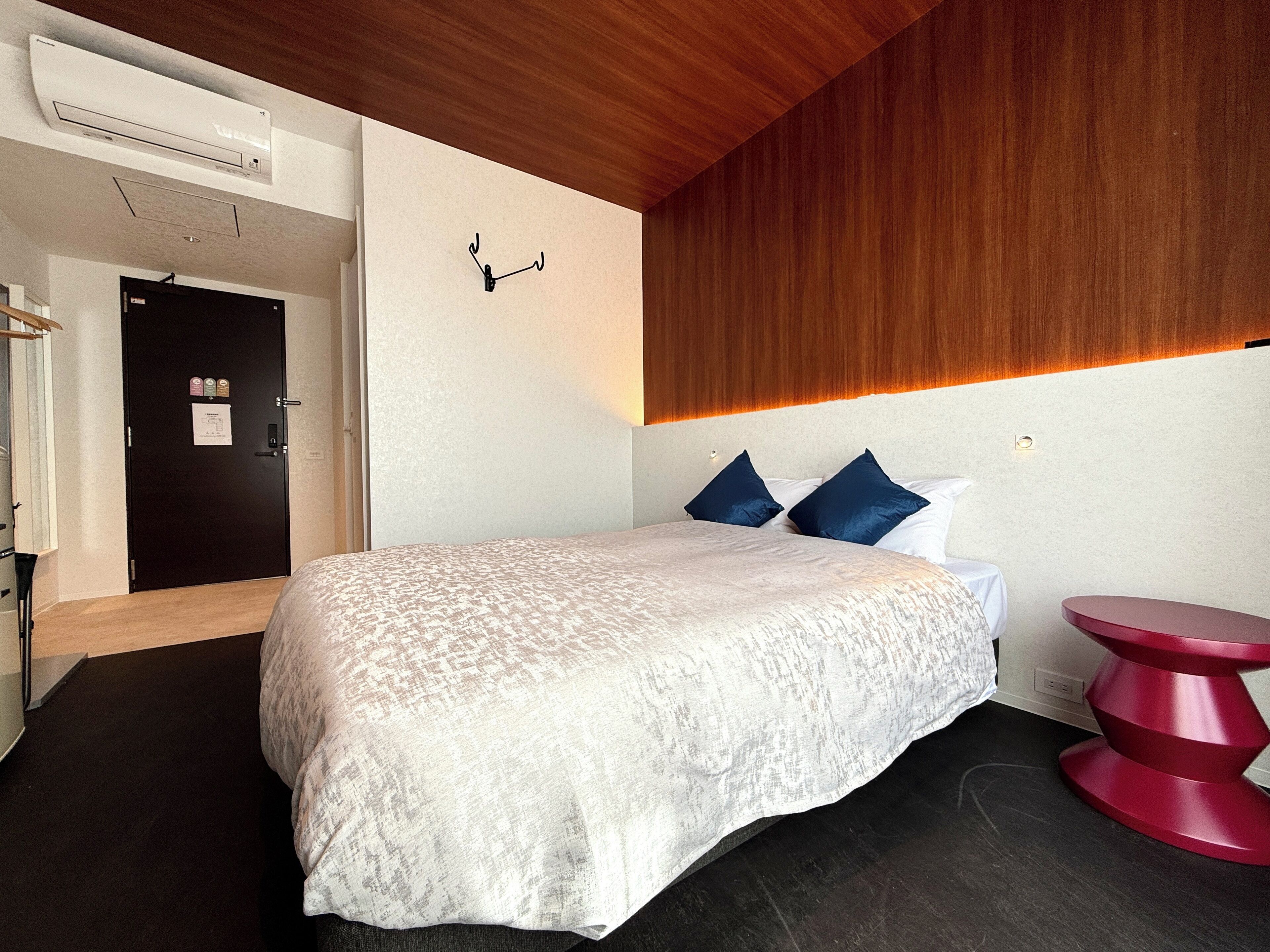 A hotel room with a bed, a wooden ceiling, a black door, a red stool, and a wall-mounted light fixture.