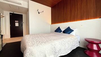 A hotel room with a bed, a wooden ceiling, a black door, a red stool, and a wall-mounted light fixture.