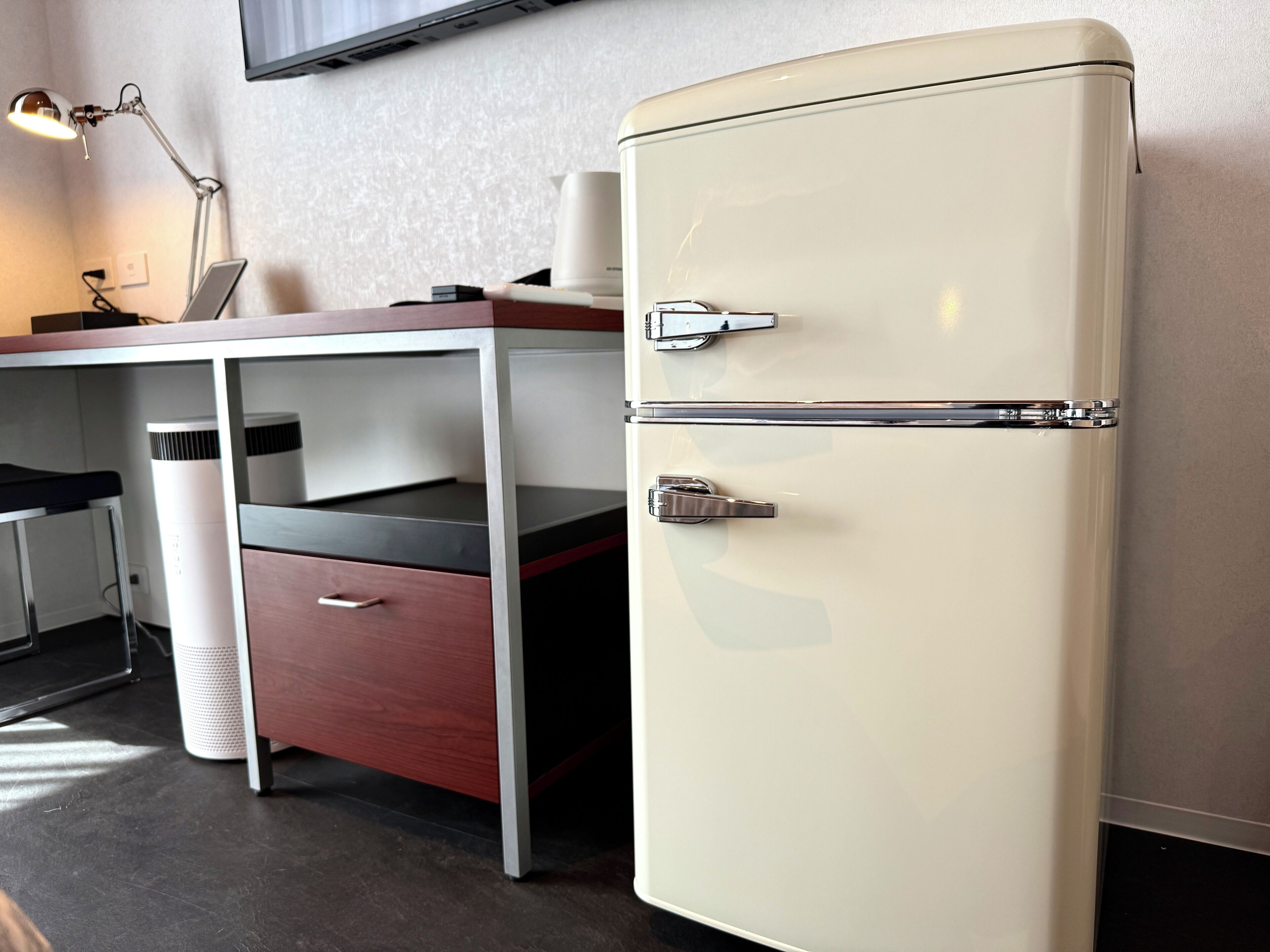 A compact kitchenette with a white refrigerator, a wooden desk, a chair, and a lamp.