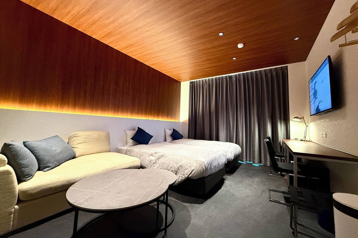 Ein modernes Hotelzimmer mit Holzbalkendecke, einer Sitzecke, einem Bett mit weißer Bettdecke, einem Schreibtisch mit Stuhl und einem flachbildfernseher an der Wand.