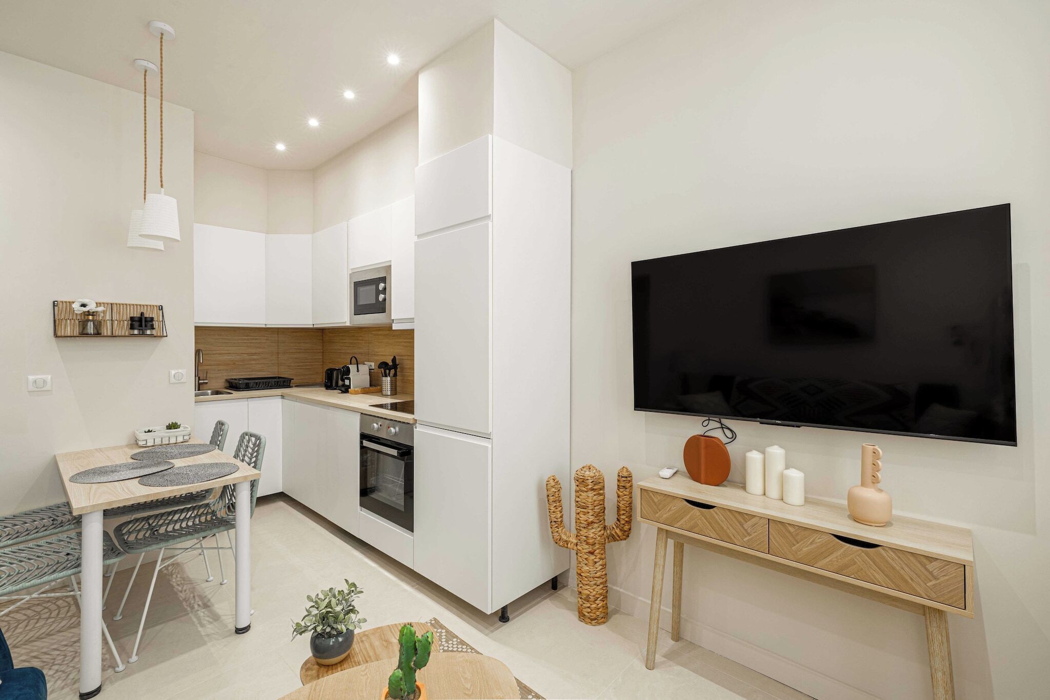 Una cocina moderna con armarios blancos, un microondas y un horno empotrado. Hay un comedor con una mesa y sillas, y un televisor montado en la pared.
