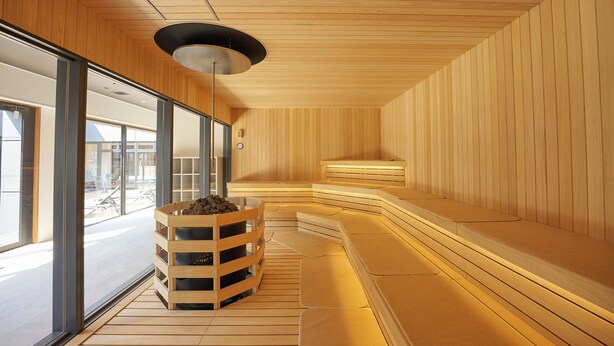 Un sauna moderne avec des murs et des bancs en bois, un foyer central et de grandes fenêtres offrant une vue sur l’extérieur.