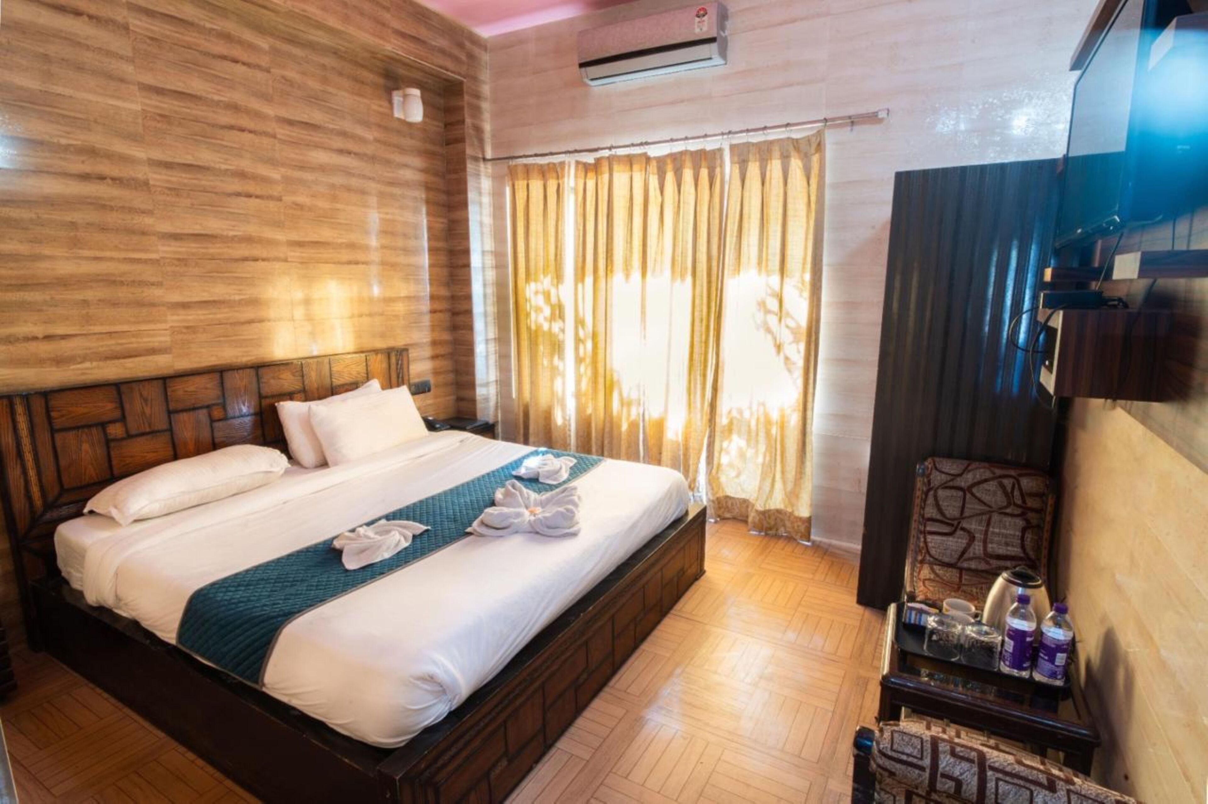 A hotel room with a wooden headboard, a bed with white linens and a teal throw, a bedside table with toiletries, and a window with curtains.