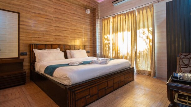 A hotel room with a wooden headboard bed, a large window with curtains, a wooden floor, and a wall-mounted air conditioner. - Sapphero Chanakya Resort  (Narendranagar)