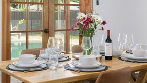 Una mesa de comedor dispuesta con platos, fuentes y copas de vino blancas, una botella de vino y un jarrón con flores.