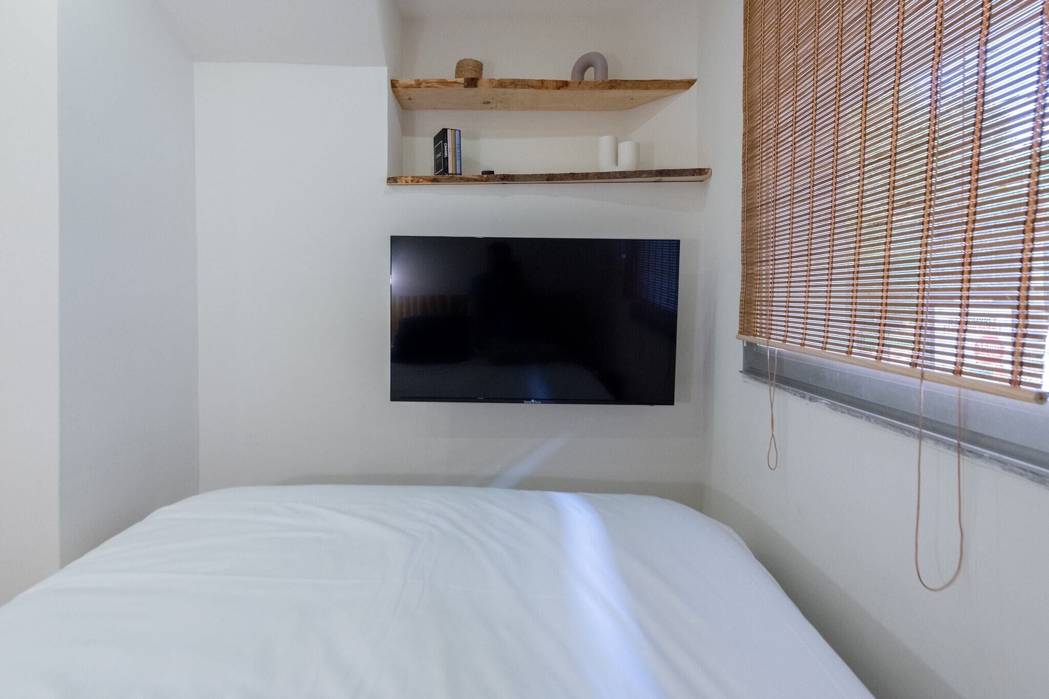 Ein Schlafzimmer mit Bett, einem an der Wand montierten Fernseher, Holzregalen und einem Fenster mit Jalousien.