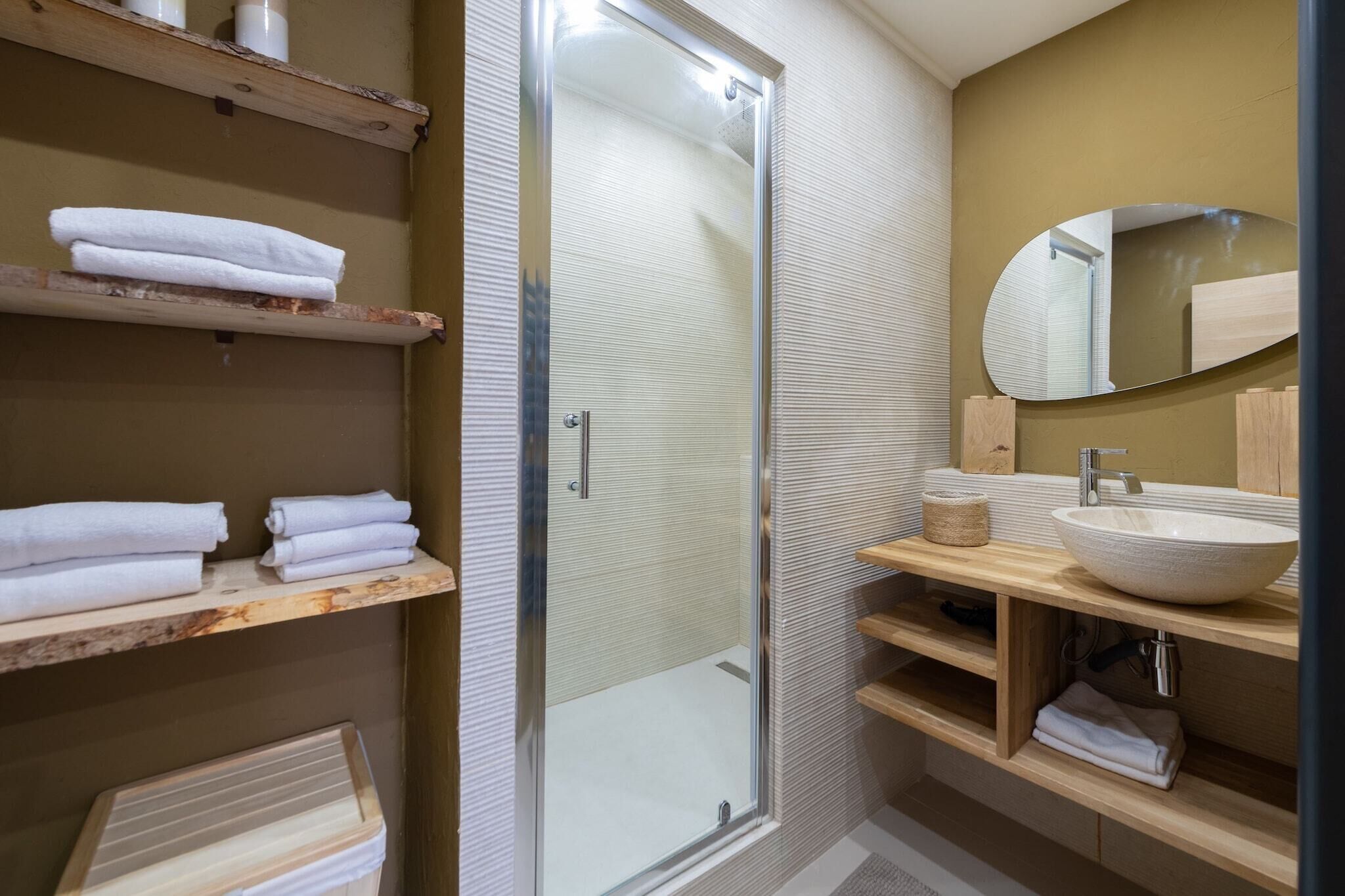 Un baño moderno con cabina de ducha de vidrio, repisas de madera con toallas, un espejo ovalado grande y un lavamanos sobre encimera.