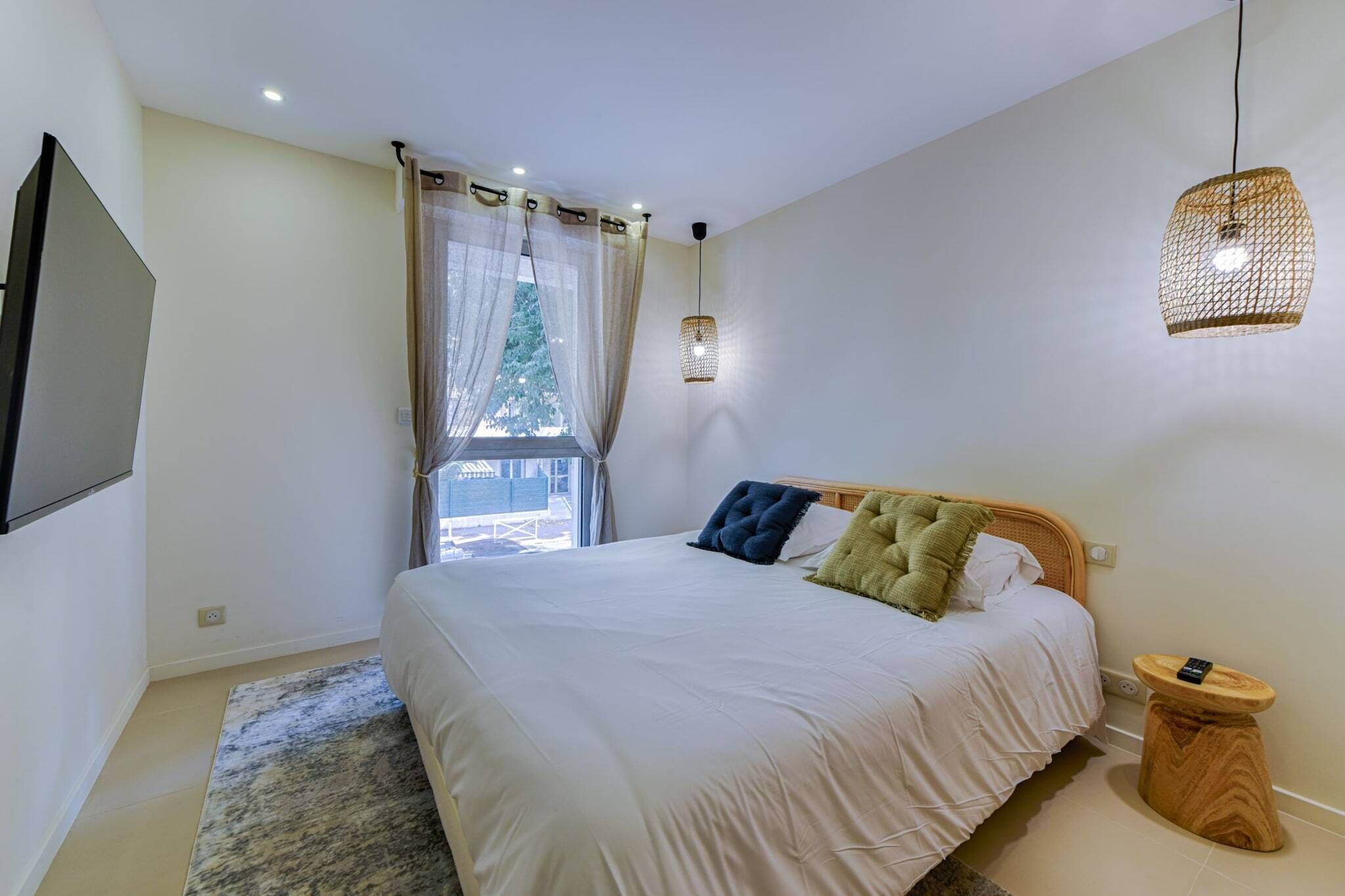 Ein Schlafzimmer mit einem Bett, einem an der Wand befestigten Fernseher, einem Fenster mit Vorhängen und einer Pendelleuchte.