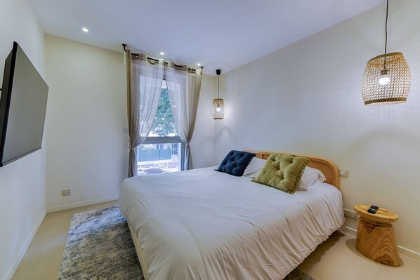 A bedroom with a bed, a TV mounted on the wall, a window with curtains, and a hanging light fixture.