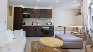 A modern living room with a white sofa, a round wicker table, and a grey ottoman. A kitchen with dark wooden cabinets and a built-in oven is visible in the background.