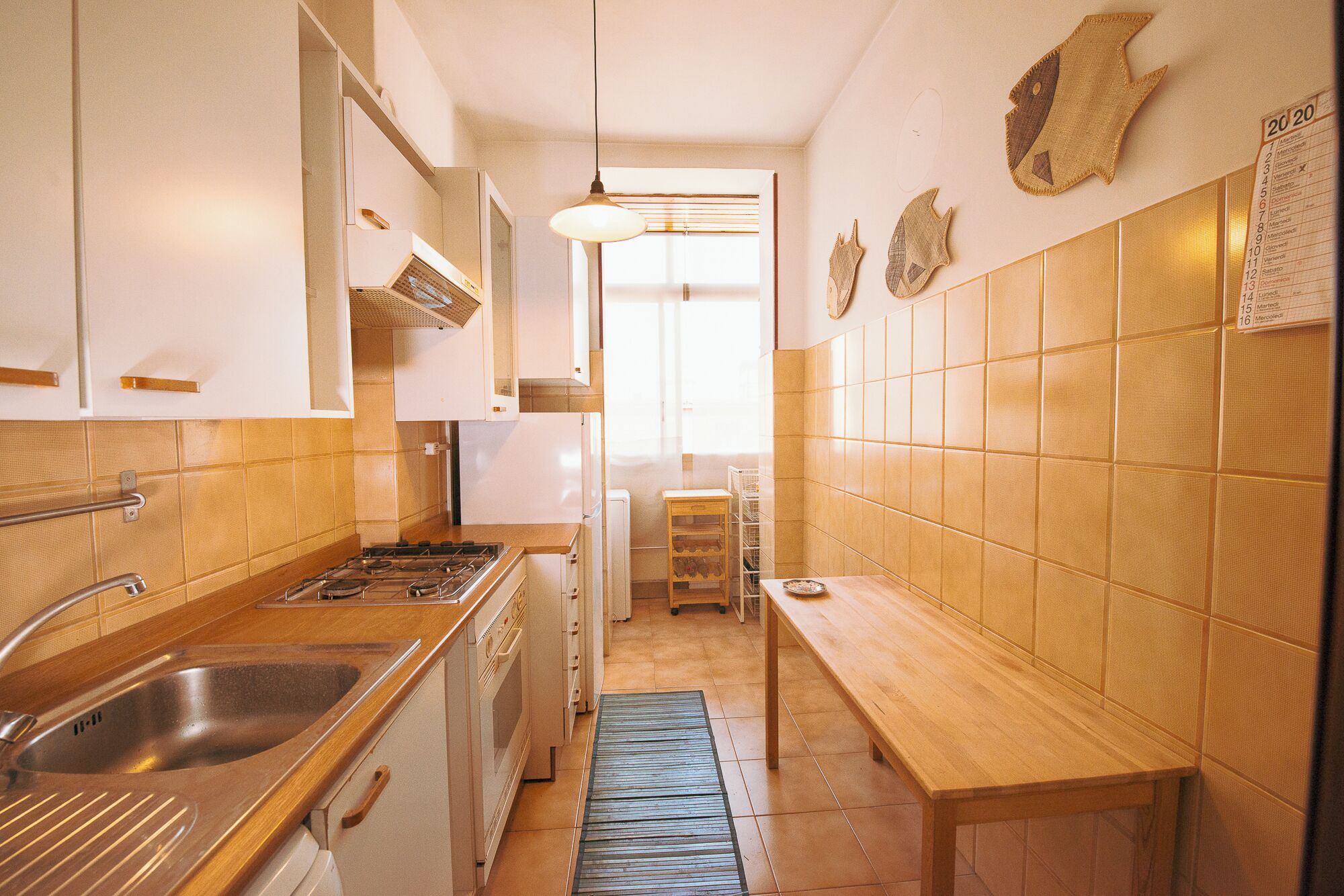 A narrow kitchen with a stove, sink, and wooden counter. There is a window with blinds and a hanging light fixture.