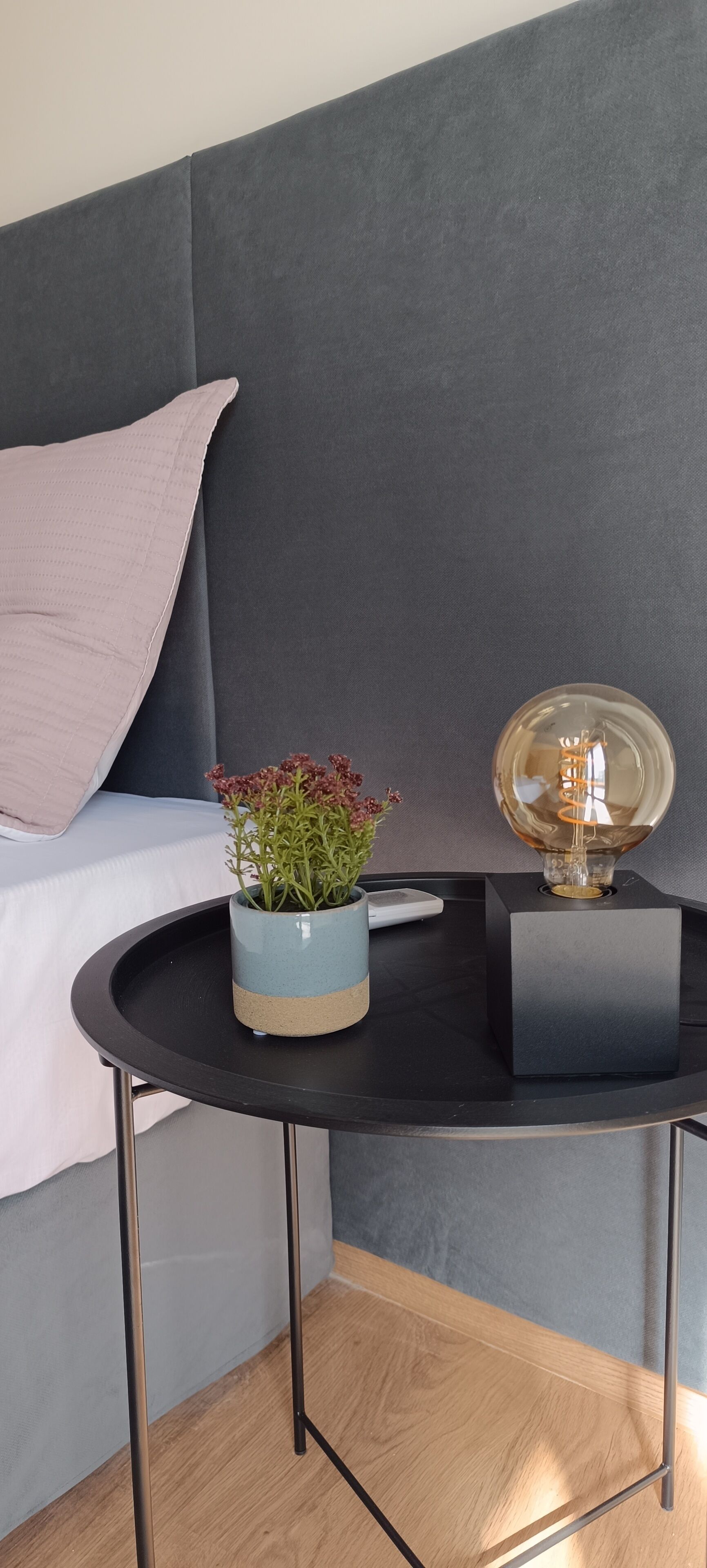 A modern bedroom with a grey headboard, a small round table with a blue vase and a decorative object, and a wooden floor.