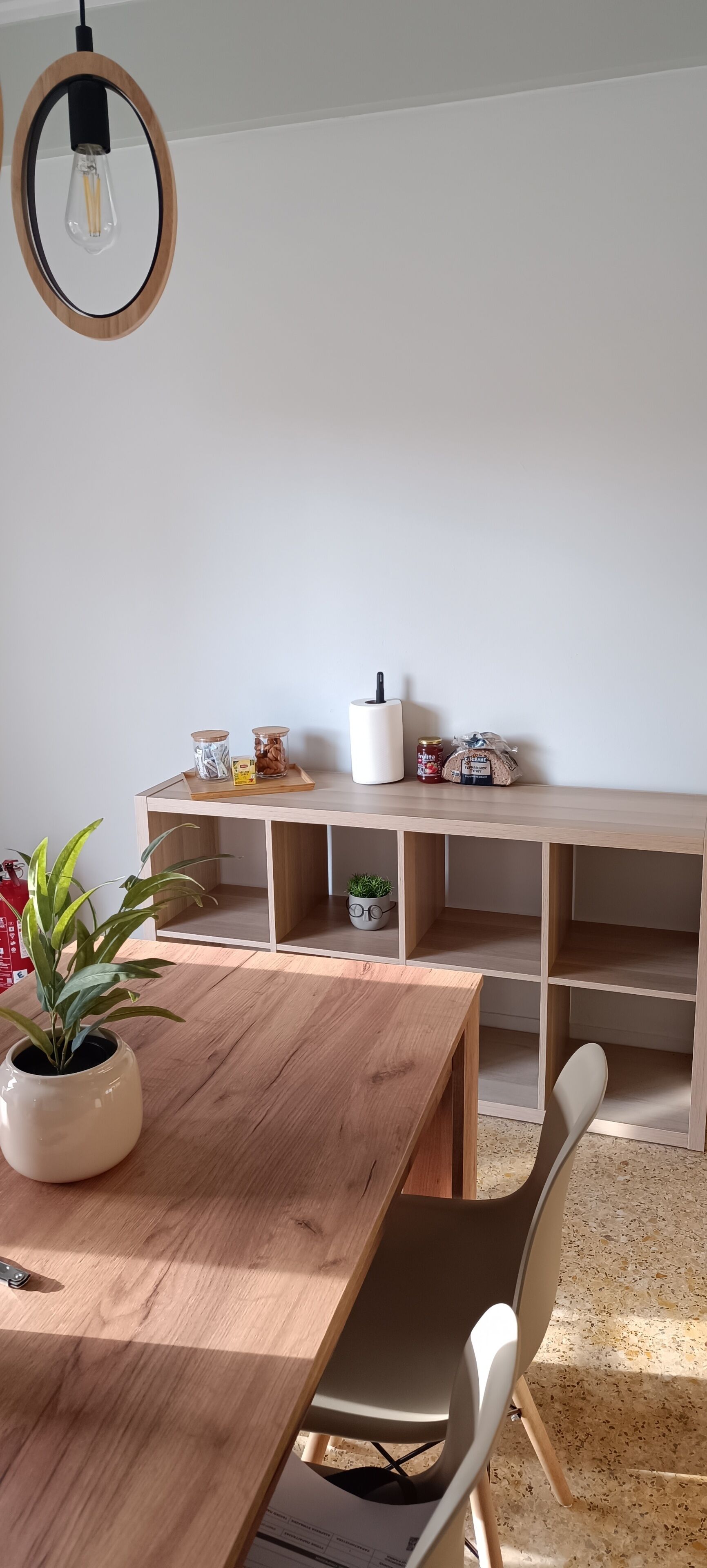 Un espace repas moderne avec une table en bois, des chaises blanches et une étagère intégrée où sont disposés des bocaux et une plante.