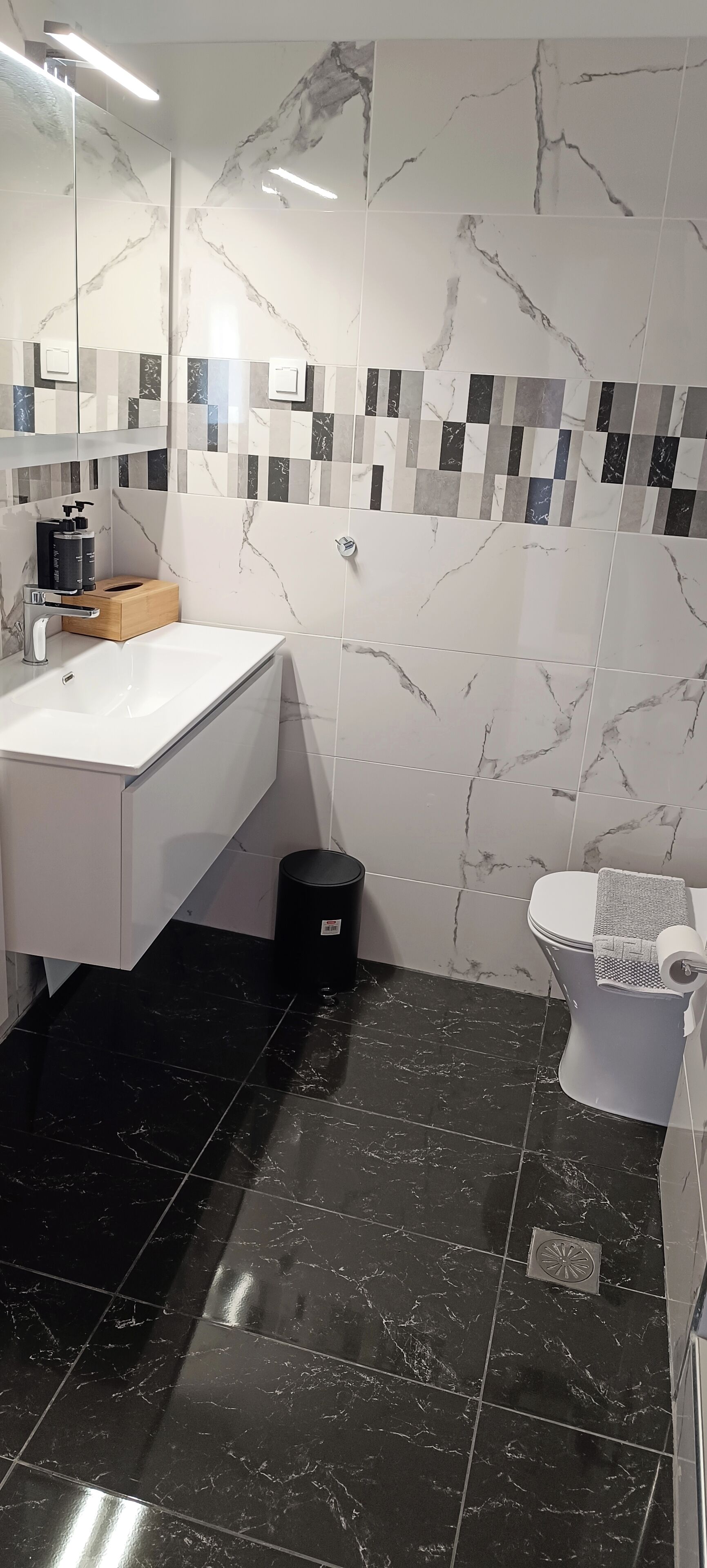 Un baño con lavamanos blanco, azulejos de mármol negro, inodoro y un cubo de basura.