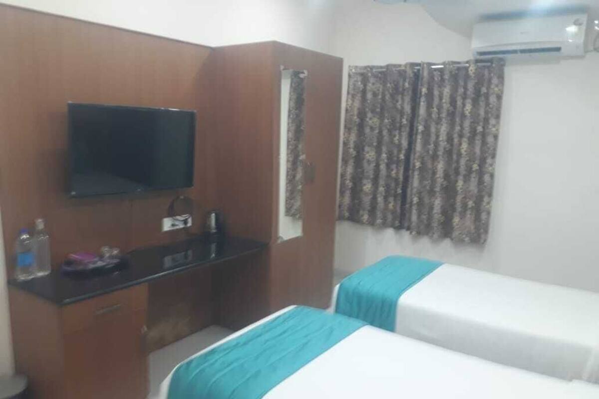 A hotel room with a flat-screen TV mounted on the wall, a wooden desk with a mirror, a small table, and two beds with blue bedspreads.