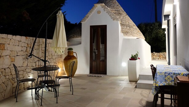 A cozy outdoor patio with stone walls, a tiled floor, a table with chairs, a large vase, and a small building with a conical roof. - Unique Trullo enchantment among olive trees (Noci)