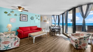 A room with a red sofa, a wooden coffee table, and two armchairs with a sea-themed pattern. There's a ceiling fan, a dining table with chairs, and a view of the ocean through large windows.